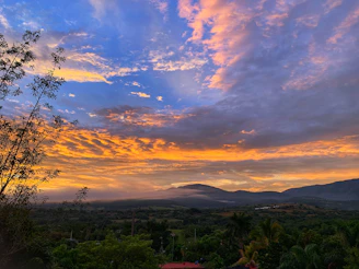 A vibrant sunset over a lush Latin American mountain landscape with colorful clouds and silhouetted trees.