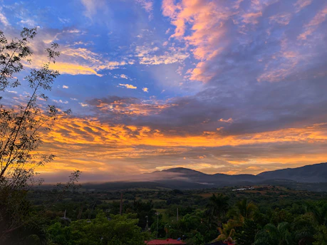 A vibrant sunset over a lush Latin American mountain landscape with colorful clouds and silhouetted trees.