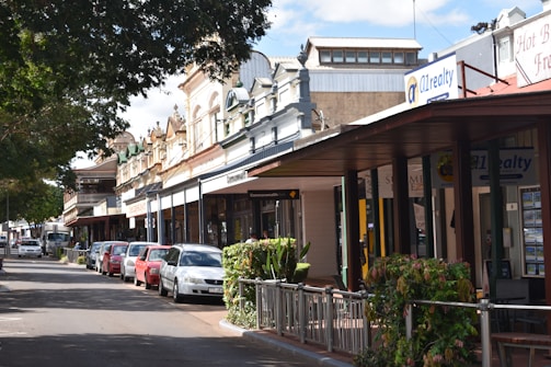 A street scene featuring a row of historic buildings with ornate facades, small businesses, and a series of parked cars along the roadside. There are trees providing shade, and a covered sidewalk with railings and plants.