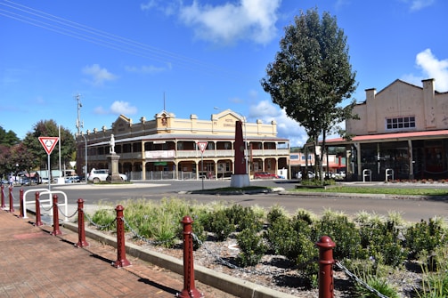 A historic-looking hotel with a two-story porch is the central feature, surrounded by a small roundabout with traffic signs. The area is well-landscaped with shrubs and trees, and an older industrial-style building is visible to the right.