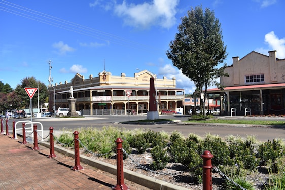 A historic-looking hotel with a two-story porch is the central feature, surrounded by a small roundabout with traffic signs. The area is well-landscaped with shrubs and trees, and an older industrial-style building is visible to the right.