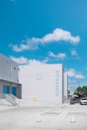 A clear blue sky with scattered clouds is visible above a modern industrial building labeled 'SUPERBLUE'. The building has a minimalist design with a light gray facade and blue accents. In the foreground, a concrete parking area with traffic cones leads to a staircase on the left side of the building. A white vehicle is parked nearby, and some greenery is seen in the distance.