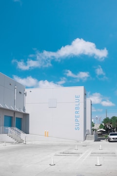 A clear blue sky with scattered clouds is visible above a modern industrial building labeled 'SUPERBLUE'. The building has a minimalist design with a light gray facade and blue accents. In the foreground, a concrete parking area with traffic cones leads to a staircase on the left side of the building. A white vehicle is parked nearby, and some greenery is seen in the distance.