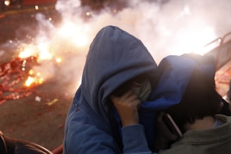 Two individuals wearing hooded jackets protect themselves from bright fireworks or explosions in the background. The scene is chaotic with smoke and scattered debris, creating a sense of urgency.