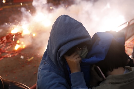 Two individuals wearing hooded jackets protect themselves from bright fireworks or explosions in the background. The scene is chaotic with smoke and scattered debris, creating a sense of urgency.