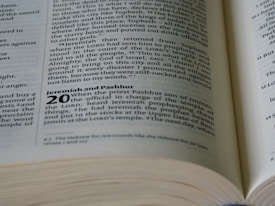 An open book displaying text from what appears to be a religious scripture, focusing on a passage titled 'Jeremiah and Pashhur'. The text is printed in black ink on cream-colored paper, with 'Jeremiah and Pashhur' in bold and larger font. The text is structured in two columns, with verse numbers highlighted.