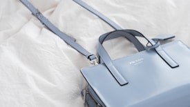 A sleek, light blue leather handbag with a detachable shoulder strap is displayed on a crumpled white fabric background. The bag features the brand name 'Prada Milano' embossed on its shiny surface.