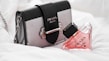 A stylish Prada handbag with a detachable chain strap sits alongside a triangular bottle of pink perfume, also branded Prada. The soft, white background enhances the luxury feel of the products.
