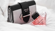 A stylish Prada handbag with a detachable chain strap sits alongside a triangular bottle of pink perfume, also branded Prada. The soft, white background enhances the luxury feel of the products.