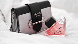A stylish Prada handbag with a detachable chain strap sits alongside a triangular bottle of pink perfume, also branded Prada. The soft, white background enhances the luxury feel of the products.