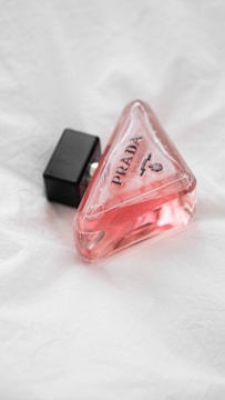 A triangular glass bottle of pink liquid with a black cap lies on a textured white surface. The bottle is labeled with 'PRADA' in bold letters.
