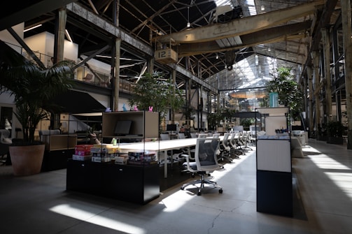 A spacious, modern office interior with high ceilings and large windows allowing natural light to illuminate the space. The area is filled with rows of white office chairs and long wooden tables. Lush green plants are strategically placed around the office, adding a touch of nature. Industrial elements such as exposed beams and metal structures are visible. Various office supplies and board games are placed on the tables.