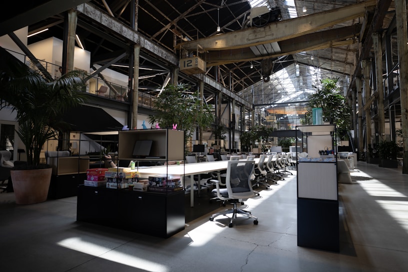 A spacious, modern office interior with high ceilings and large windows allowing natural light to illuminate the space. The area is filled with rows of white office chairs and long wooden tables. Lush green plants are strategically placed around the office, adding a touch of nature. Industrial elements such as exposed beams and metal structures are visible. Various office supplies and board games are placed on the tables.