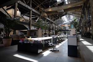 A spacious, modern office interior with high ceilings and large windows allowing natural light to illuminate the space. The area is filled with rows of white office chairs and long wooden tables. Lush green plants are strategically placed around the office, adding a touch of nature. Industrial elements such as exposed beams and metal structures are visible. Various office supplies and board games are placed on the tables.
