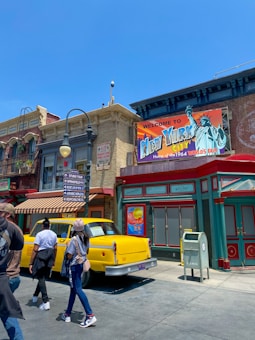 A vibrant street scene with a New York City theme, featuring a prominent yellow taxi and colorful storefronts. The buildings have classic architectural designs with a vintage vibe. A large sign welcomes visitors to New York City, highlighting the 1964 World's Fair. Several people are walking by, adding a lively atmosphere.