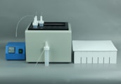 A laboratory apparatus setup featuring a white machine with two nozzles and tubes on top, a blue control unit with digital display and buttons, and a white rectangular box with slotted openings.