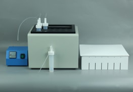 A laboratory apparatus setup featuring a white machine with two nozzles and tubes on top, a blue control unit with digital display and buttons, and a white rectangular box with slotted openings.