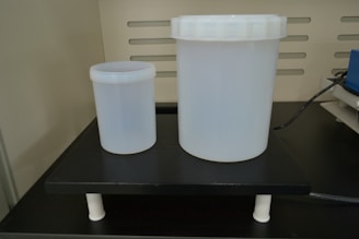 Two translucent plastic containers of different sizes are placed on a black laboratory platform. The larger container has a screw-top lid, and both are cylindrical. The background features vertical slits and part of electronic equipment with a connecting cable.