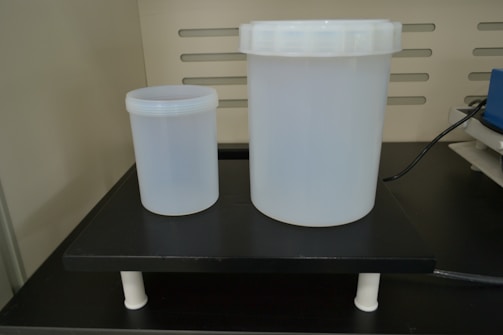 Two translucent plastic containers of different sizes are placed on a black laboratory platform. The larger container has a screw-top lid, and both are cylindrical. The background features vertical slits and part of electronic equipment with a connecting cable.