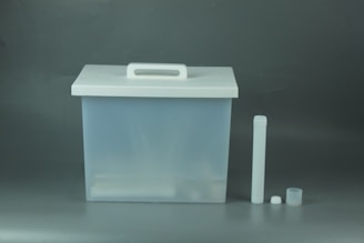 A translucent plastic storage container with a white lid featuring a handle is positioned on the left side. To the right of the container are a long cylindrical tube and two smaller cylindrical objects.
