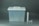 A translucent plastic storage container with a white lid featuring a handle is positioned on the left side. To the right of the container are a long cylindrical tube and two smaller cylindrical objects.