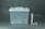 A translucent plastic storage container with a white lid featuring a handle is positioned on the left side. To the right of the container are a long cylindrical tube and two smaller cylindrical objects.
