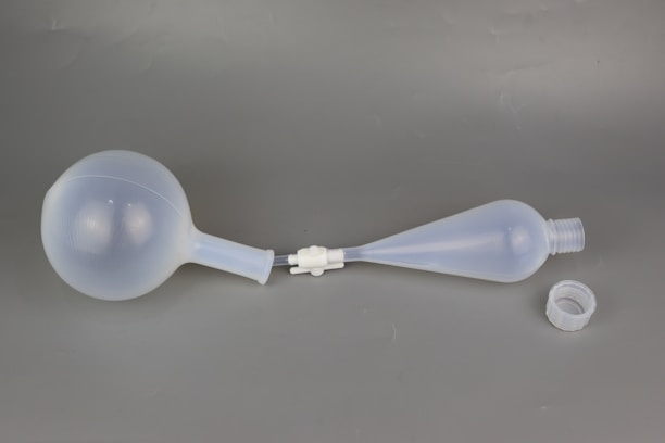 A laboratory glassware item consisting of two connected bulbs with a narrow neck, lying on a neutral background. The larger bulb has a spherical shape, while the smaller one tapers into a conical form with an open top and a detachable screw cap placed beside it.