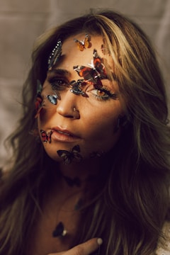 A woman gazes intently, adorned with colorful butterfly stickers across her face, creating an artistic and surreal effect. Her hair cascades around her shoulders, highlighted by soft lighting that adds warmth to the scene.