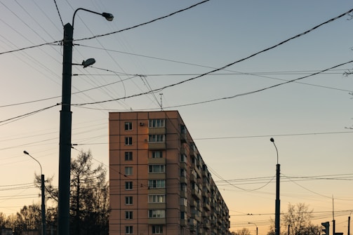 A residential building with multiple floors is situated alongside several utility poles. Numerous power lines crisscross the scene, stretching across the vivid evening sky with faint pink and blue hues. Trees without leaves are visible in the foreground and background, adding an urban and slightly stark atmosphere.