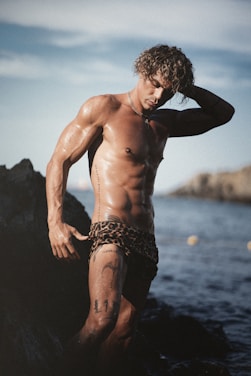 A muscular man with curly hair poses near a rocky shoreline, wearing leopard print swimwear. His skin glistens, possibly from water or sweat, and he has several tattoos. The background reveals a calm sea and distant rocks with a clear blue sky overhead.