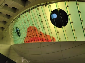 A large, decorated panel with colored glass segments featuring an abstract design. The artwork displays an orange-red geometric shape surrounded by green and yellow hues, along with dark circular elements resembling eyes.