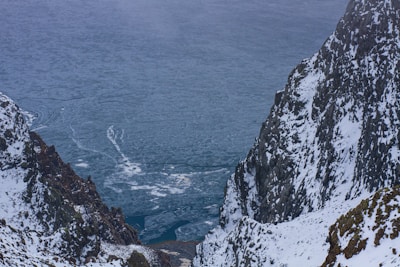 A frozen lake is surrounded by rugged snow-covered mountains. The icy surface of the water features cracks and various patterns. The scene contrasts the dark, jagged rock formations against the pristine whiteness of the snow.