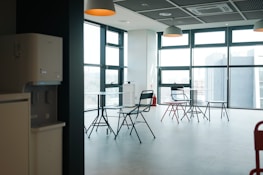 A modern and minimalist office space with large windows allowing for ample natural light. The room contains several black and red folding chairs arranged around small round tables. The ceiling has grid-like panels with hanging pendant lights. A water dispenser and air conditioning unit are visible against the wall. The overall atmosphere is clean and spacious.