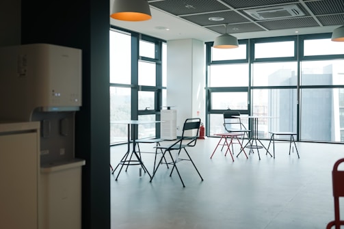 A modern and minimalist office space with large windows allowing for ample natural light. The room contains several black and red folding chairs arranged around small round tables. The ceiling has grid-like panels with hanging pendant lights. A water dispenser and air conditioning unit are visible against the wall. The overall atmosphere is clean and spacious.