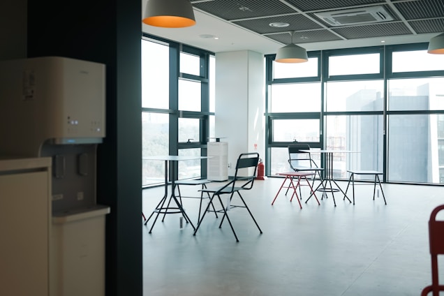 A modern and minimalist office space with large windows allowing for ample natural light. The room contains several black and red folding chairs arranged around small round tables. The ceiling has grid-like panels with hanging pendant lights. A water dispenser and air conditioning unit are visible against the wall. The overall atmosphere is clean and spacious.
