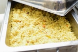 A large stainless steel serving tray filled with cooked rice mixed with small pieces of vegetables like carrots and peas.