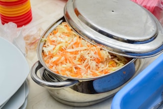 A stainless steel pot filled with shredded carrots and cabbage sits on a table. The lid is partially covering the pot. In the background, there are stacks of colorful plastic plates and a blue container.