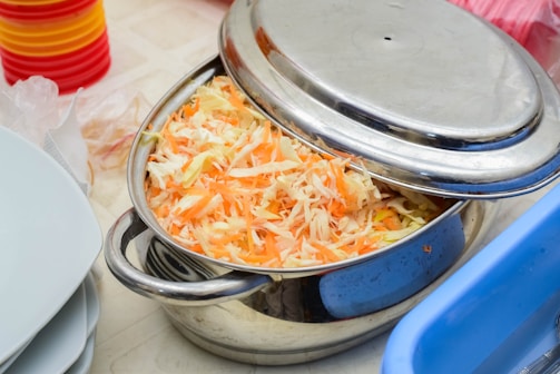 A stainless steel pot filled with shredded carrots and cabbage sits on a table. The lid is partially covering the pot. In the background, there are stacks of colorful plastic plates and a blue container.