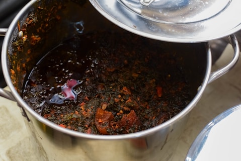 A stainless steel pot containing a rich, dark-colored stew with visible pieces of vegetables or herbs and chunks of what seem to be meat. The surface is glossy, indicating the presence of oils or broth. The pot lid is partially open, reflecting light.