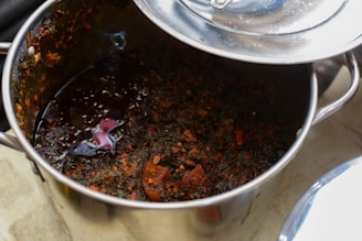 A stainless steel pot containing a rich, dark-colored stew with visible pieces of vegetables or herbs and chunks of what seem to be meat. The surface is glossy, indicating the presence of oils or broth. The pot lid is partially open, reflecting light.