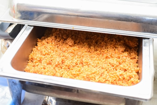 A large metal chafing dish containing orange-colored rice, likely seasoned or spiced, with a stainless steel lid that is partially open.