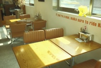 A small dining area with wooden tables and brown padded chairs, situated near a window. Sunlight is streaming in, casting shadows on the tables. There is a decorative item resembling a smiling yellow square on the windowsill. The wall has an orange text message. Table number two has a napkin holder and menu. The overall setting is calm and neatly arranged.