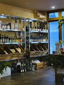 A cozy interior with wooden shelves holding a variety of wine bottles and packaged goods. Glass doors allow natural light to enter, and several plants add a touch of greenery. There are wooden boxes on the floor filled with additional items.