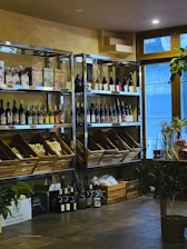A cozy interior with wooden shelves holding a variety of wine bottles and packaged goods. Glass doors allow natural light to enter, and several plants add a touch of greenery. There are wooden boxes on the floor filled with additional items.