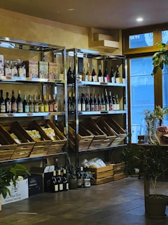A cozy interior with wooden shelves holding a variety of wine bottles and packaged goods. Glass doors allow natural light to enter, and several plants add a touch of greenery. There are wooden boxes on the floor filled with additional items.