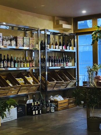 A cozy interior with wooden shelves holding a variety of wine bottles and packaged goods. Glass doors allow natural light to enter, and several plants add a touch of greenery. There are wooden boxes on the floor filled with additional items.