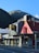 A McDonald's and KFC restaurant sit side by side on a street corner with a mountainous landscape in the background. The McDonald's features a modern black and gray exterior, while the KFC has a red peaked roof with the Colonel Sanders logo. The establishments are located on Cow Lane, as indicated by the street sign. The scene is well lit by sunlight, casting shadows on the pavement.