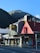 A McDonald's and KFC restaurant sit side by side on a street corner with a mountainous landscape in the background. The McDonald's features a modern black and gray exterior, while the KFC has a red peaked roof with the Colonel Sanders logo. The establishments are located on Cow Lane, as indicated by the street sign. The scene is well lit by sunlight, casting shadows on the pavement.