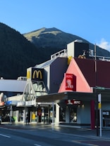 A McDonald's and KFC restaurant sit side by side on a street corner with a mountainous landscape in the background. The McDonald's features a modern black and gray exterior, while the KFC has a red peaked roof with the Colonel Sanders logo. The establishments are located on Cow Lane, as indicated by the street sign. The scene is well lit by sunlight, casting shadows on the pavement.