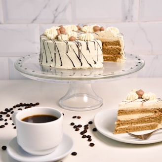 A layered cake with cream and chocolate drizzle is displayed on a glass stand. The cake is decorated with dollops of cream and small round chocolates on top. A slice of the cake is served on a plate with a fork, and a cup of black coffee sits nearby. Coffee beans are scattered on the white surface, adding contrast.